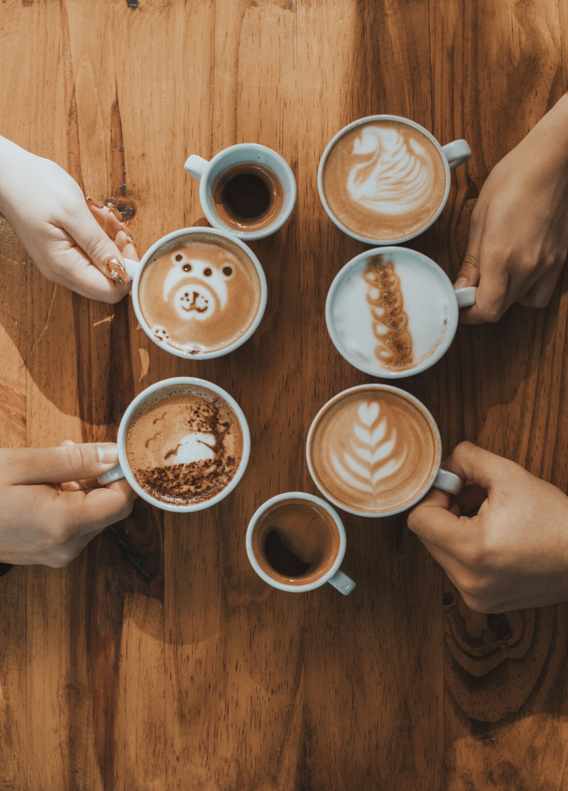 A creative display of latte art in a cafe, showing diverse coffee designs held by hands on a wooden table.